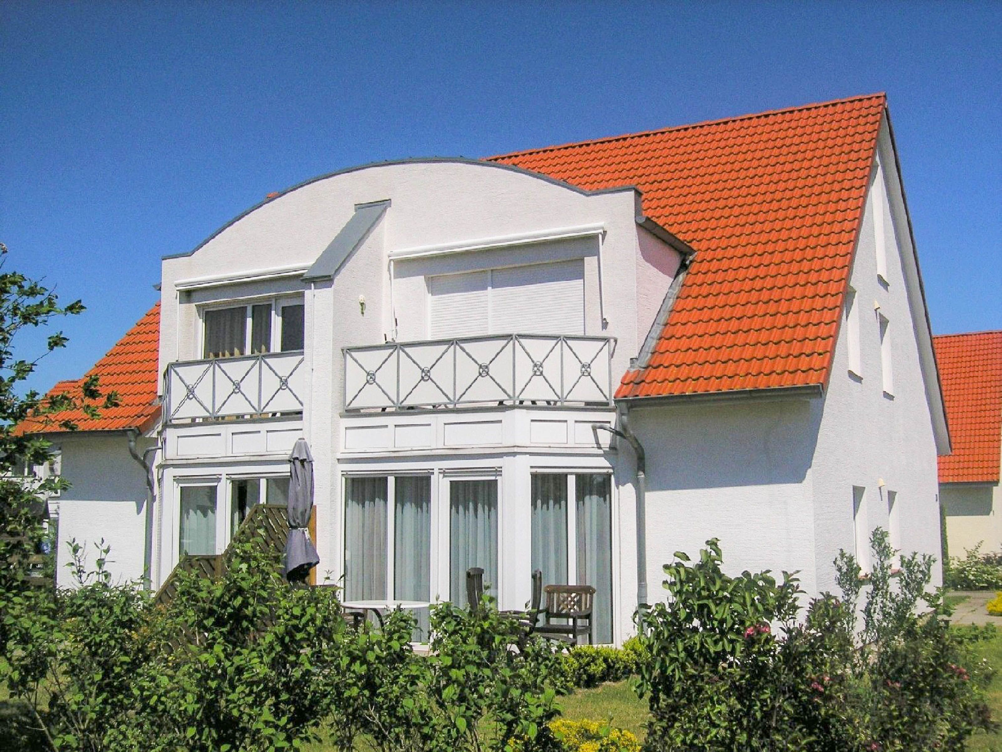 Une maison à deux étages, avec un toit recouvert de tuiles rouges, des murs extérieurs blancs et un balcon doté d’une balustrade blanche.