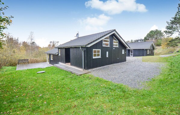 A black wooden building with a sloped roof, a gravel driveway, and a grassy area with a bench.