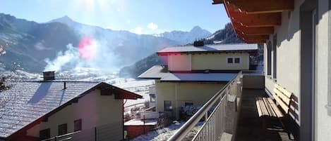 A mountain view from a balcony with a metal railing, a wooden bench, and a building with a red roof.