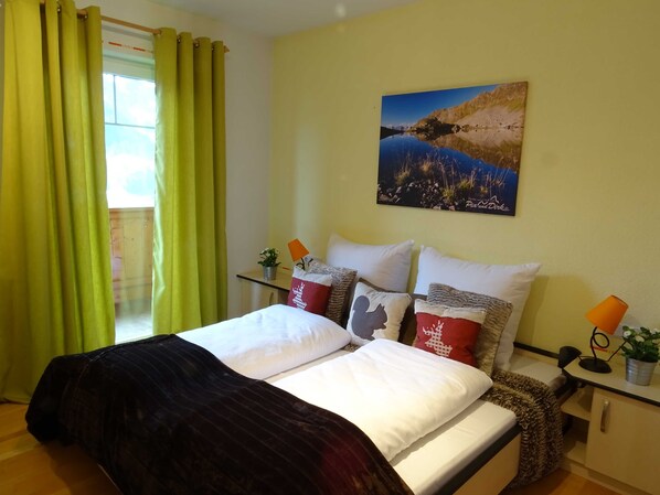 A hotel room with a bed, pillows, a nightstand, a lamp, a window with curtains, and a painting on the wall.