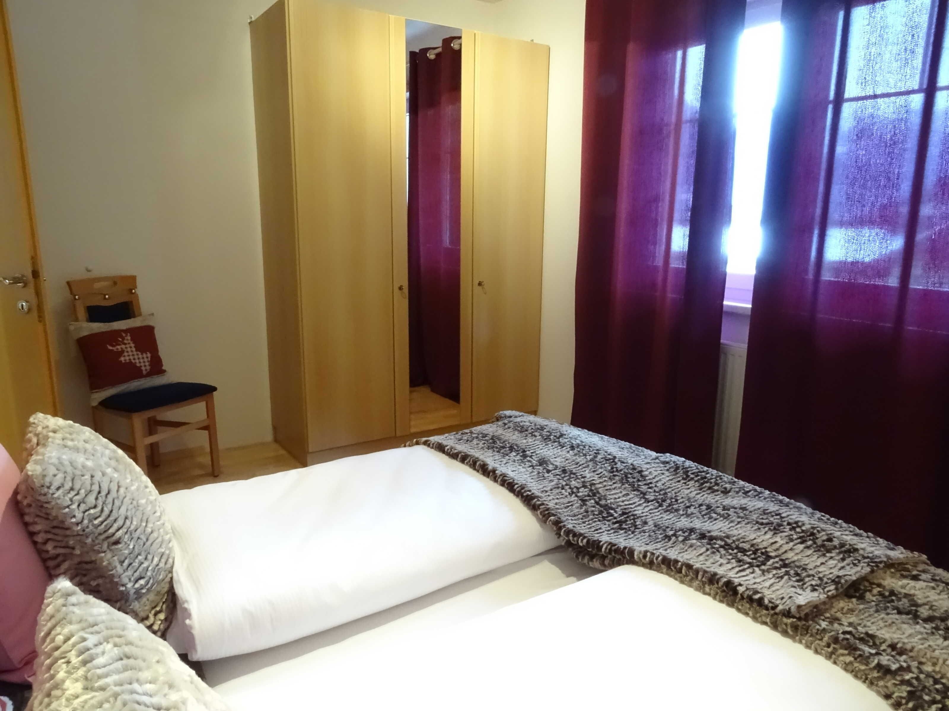 A bedroom with a bed, a wardrobe, a chair, and a window with curtains.