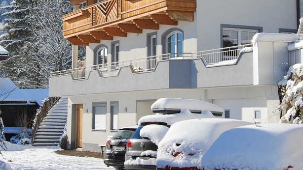 Ein mehrstöckiges Gebäude mit einem hölzernen Balkon und einem schneebedeckten Berg im Hintergrund, sowie abgestellte Autos.
