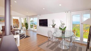 Ein modernes Wohnzimmer mit einem Glastisch, transparenten Stühlen, einem Flachbildfernseher und großen Fenstern mit herrlicher Aussicht.