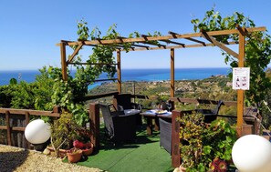 Relax in a shaded outdoor space with a pergola, wicker chairs, and a dining table, surrounded by lush greenery and a scenic coastal view.