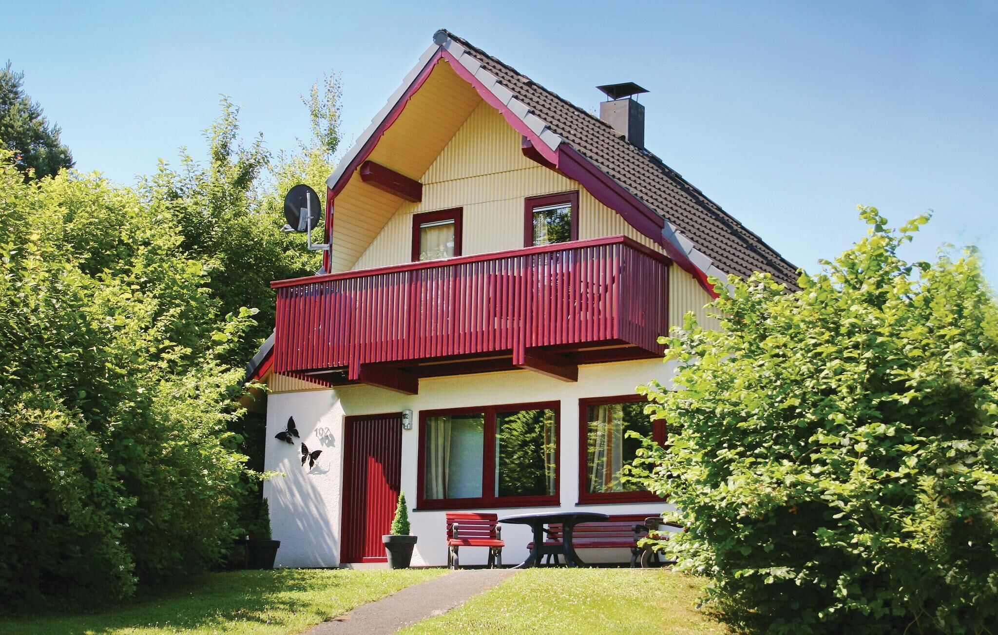 Een huis met twee verdiepingen, met een rood balkon, een kleine tuin en een pad dat naar een bankje leidt.