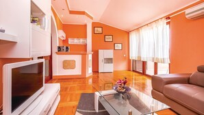 Ein modernes Wohnzimmer mit Flachbildfernseher, einer Couch, einem Glastisch und einer Küchenzeile inklusive Schränken und Kühlschrank.