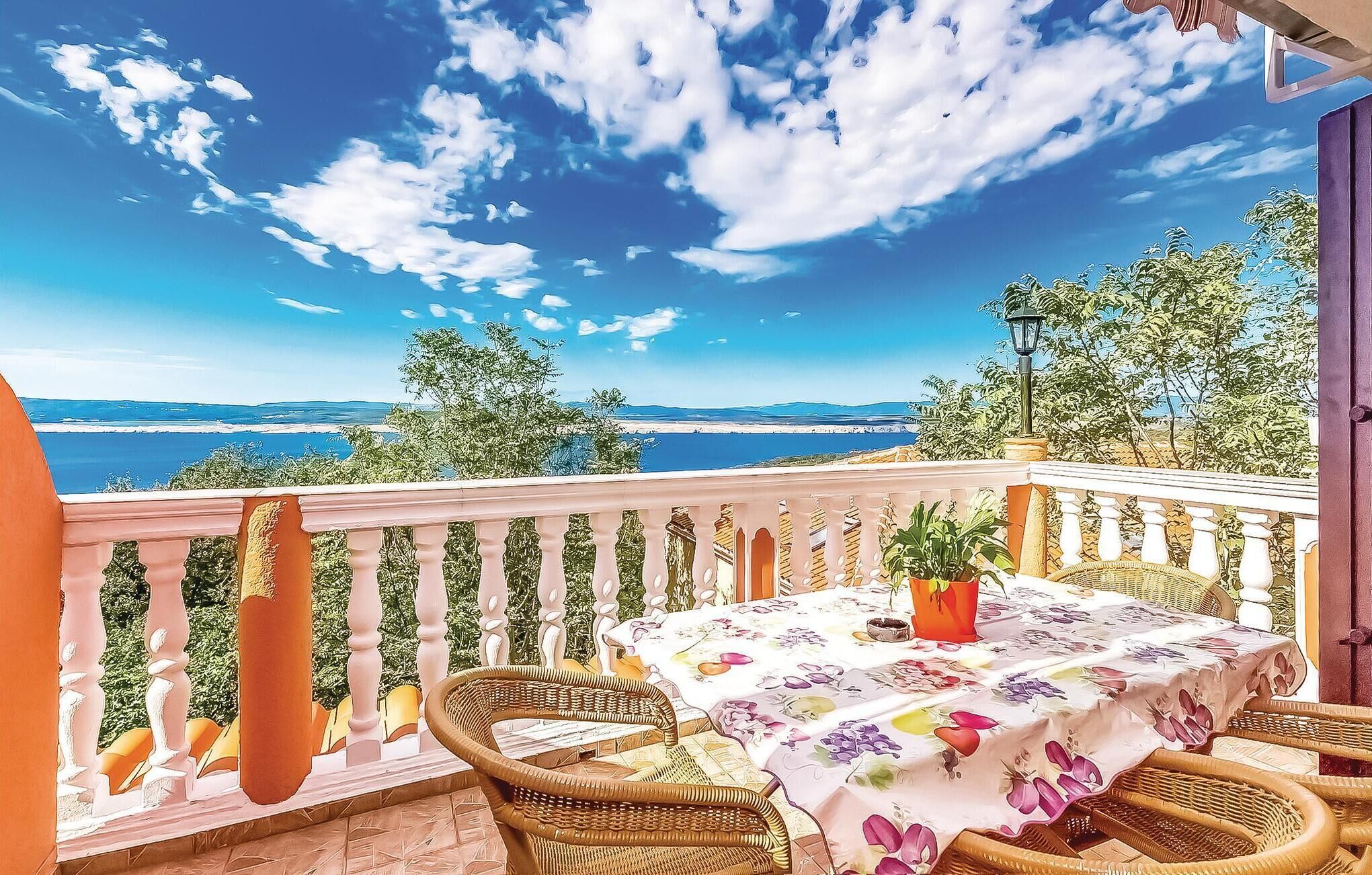 Un balcón con sillas de mimbre, una mesa con mantel floreado y vistas al mar y a las montañas.