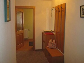 Ein schmaler Flur mit einer Holztür, einem Heizkörper, einer Bank mit einem roten Gegenstand und einem Schrank aus Holz mit Haken.