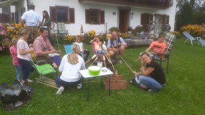 Un groupe de personnes s’est rassemblé autour d’un feu de camp dans un espace extérieur verdoyant, avec une maison en arrière-plan.