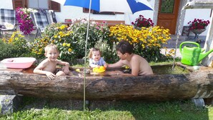 Kinder spielen in einem baumstammförmigen Wasserspiel mit Garten und einem Gebäude im Hintergrund.