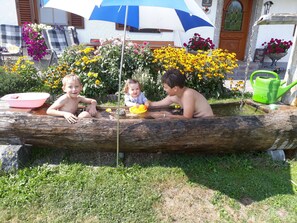 Des enfants jouent dans une fontaine d’eau en forme de tronc d’arbre, avec un jardin et un bâtiment en arrière-plan.