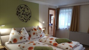 A double bed with floral bedding and multiple pillows, a bedside table, a wall decoration, a window with curtains, and a door leading to another room. - Apartment 1 - Ferienhof Nussbaumer (Gaisberg)