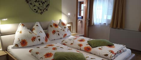 A double bed with floral bedding and multiple pillows, a bedside table, a wall decoration, a window with curtains, and a door leading to another room.