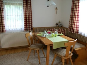 Un coin repas avec une table en bois, des chaises, un banc, un radiateur, une croix accrochée au mur et des rideaux.