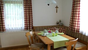 A dining area with a wooden table, chairs, a bench, a radiator, a cross on the wall, and curtains. - Apartment 1 - Ferienhof Nussbaumer (Gaisberg)