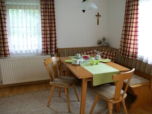 A dining area with a wooden table, chairs, a bench, a radiator, a cross on the wall, and curtains.