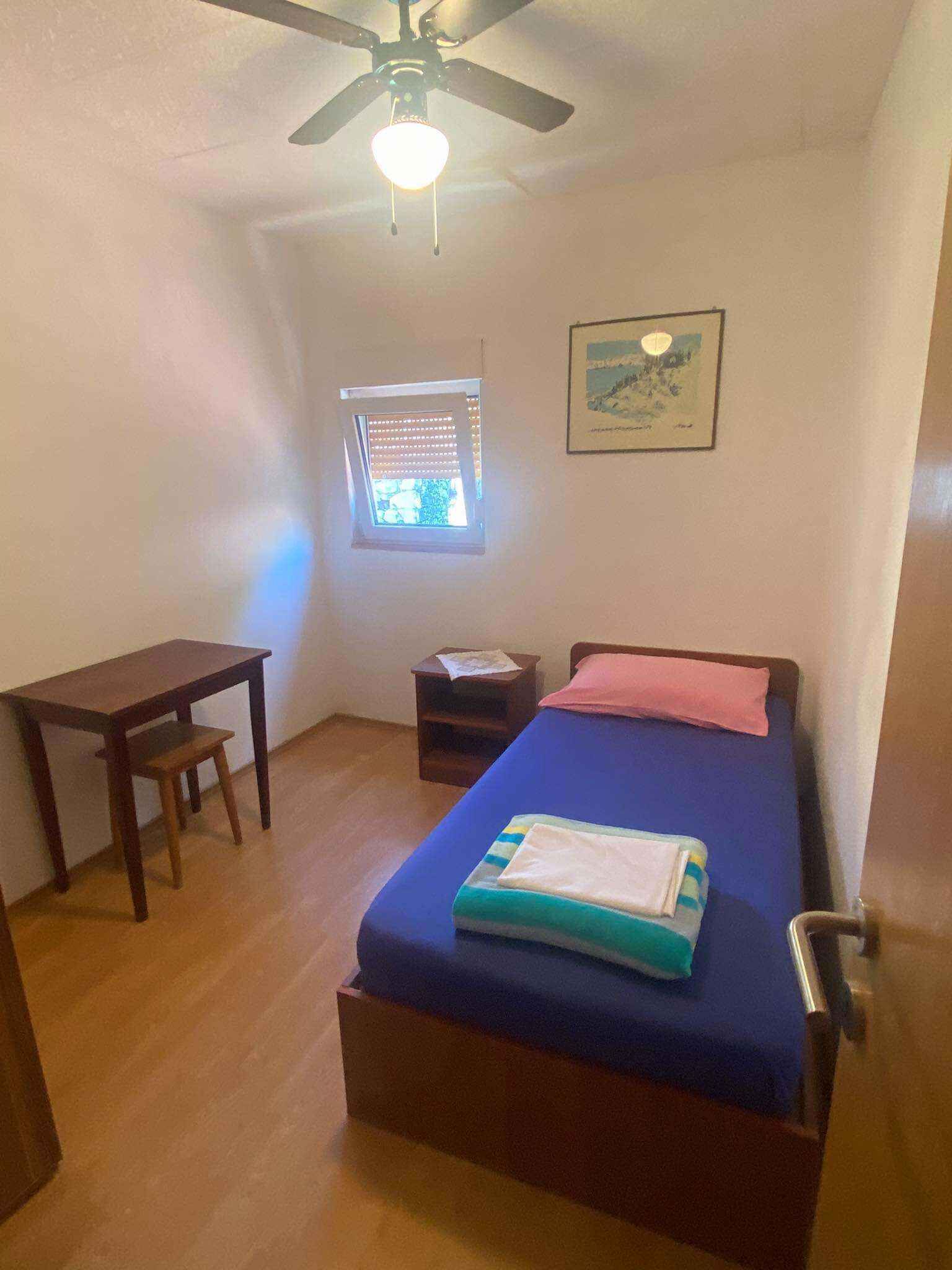 Ein kleiner, gut beleuchteter Raum mit einem Bett, einem Schreibtisch, einem Stuhl, einem Fenster mit Jalousien und einem Deckenventilator.