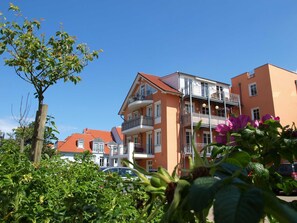 Um edifício residencial de vários andares, com varandas, telhado de telhas vermelhas e céu azul claro.
