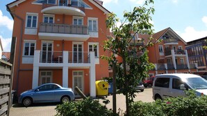 Un immeuble résidentiel à plusieurs étages, doté de balcons, avec une voiture garée devant et un arbre au premier plan.
