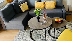 Una sala moderna con un sofá gris, cojines amarillos, una mesa de centro redonda de madera y una mesita auxiliar con un jarrón de flores y una botella.
