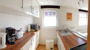 A compact kitchen with white cabinets, a wooden countertop, a coffee maker, a toaster, a microwave, a sink, and a window with a view of buildings. - Apartment 03.12 - House 12 (Trassenheide)