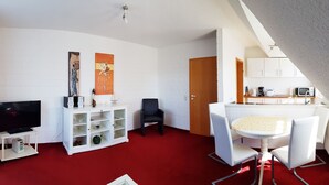 A compact living space with a TV, white entertainment unit, red carpet, dining area with white chairs, and a kitchen with white cabinets. - Apartment 03.12 - House 12 (Trassenheide)