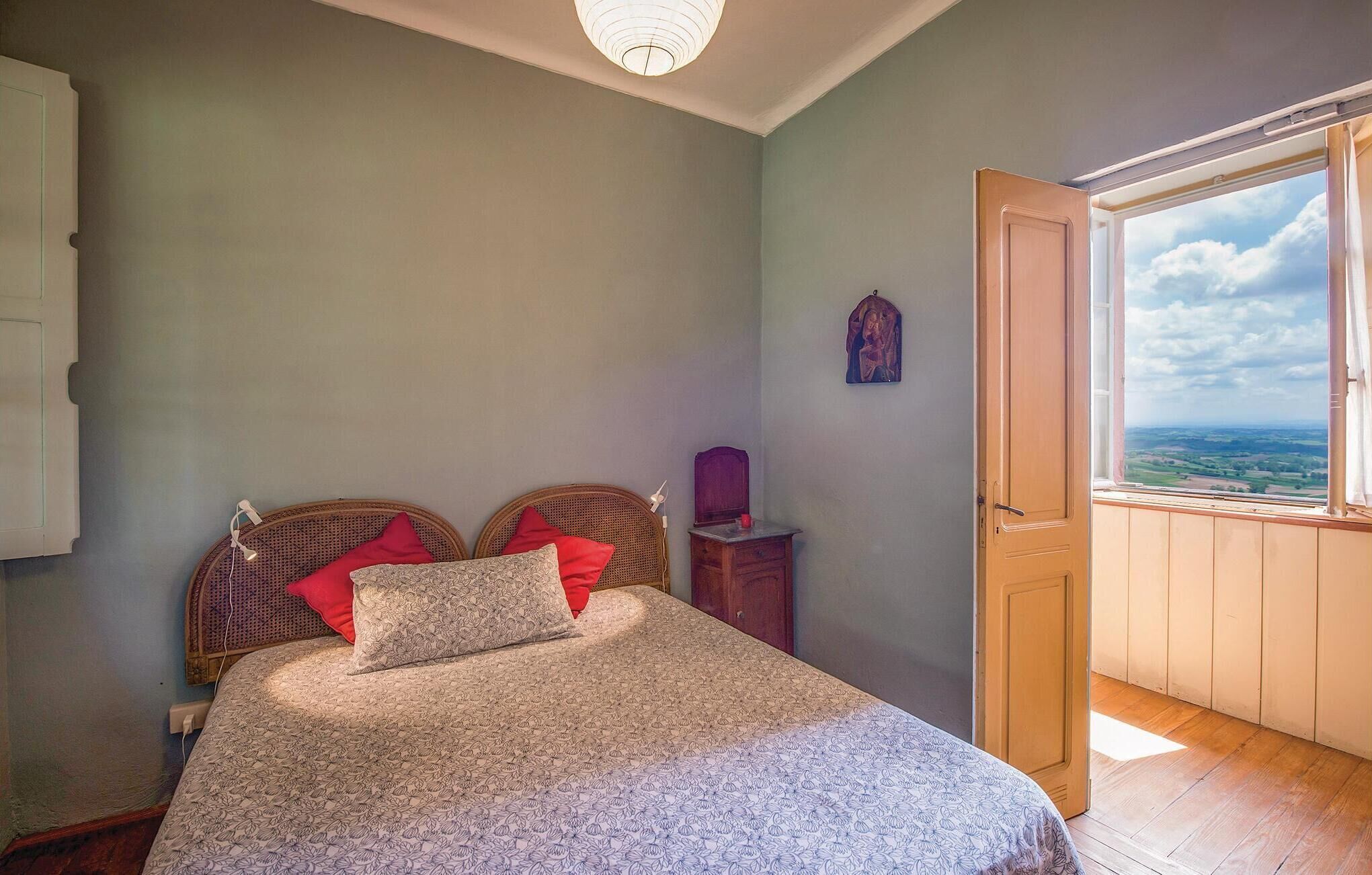 Ein Schlafzimmer mit einem Bett, zwei roten Kissen, einem Nachttisch, einem kleinen Tisch, einem Wandbild und einer Tür zu einem Balkon mit Blick.