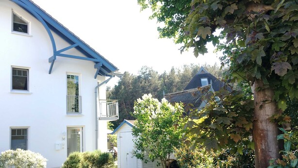 Een wit gebouw met blauwe accenten, een klein balkon en een tuin met verschillende planten en een boom.