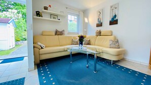Een woonkamer met een beige bank, een glazen salontafel en een blauw tapijt met een patroon. Er staan planken met decoratieve objecten en een raam met gordijnen.