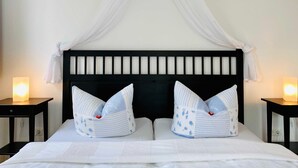 Een bed met een zwart hoofdeinde, twee kussens met bloemendesign, een witte hemel en bedlampjes.