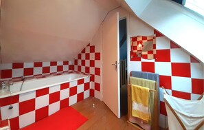 Un baño pequeño y funcional con un revestimiento de baldosas blancas y rojas a cuadros, una bañera blanca y suelo de madera.