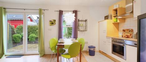 A modern kitchen with a dining area, featuring a large window, sliding doors, and a small outdoor garden visible through the window.