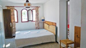 Un dormitorio con una cama, una silla de madera, una mesita de noche, una lámpara y un ventanal con cortinas.