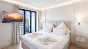 A hotel room with a bed, a TV on the wall, a balcony with curtains, and a nightstand with a lamp. - Amazing apartment in Binz with house sea view (Prora/Rügen)