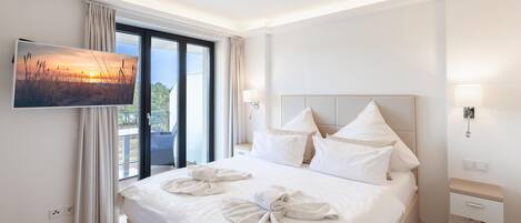 Habitación de hotel con cama, televisor en la pared, balcón con cortinas y mesita de noche con lámpara.