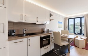 A modern kitchen with white cabinets, a wooden countertop, and a built-in oven. There is a dining area with a table and chairs, and a large window offering a view of trees. - Amazing apartment in Binz with house sea view (Prora/Rügen)