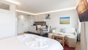 Ett hotellrum med en säng, en soffa, ett litet bord, ett litet kök och en tavla på väggen.