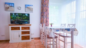 Un coin repas avec une table et des chaises blanches, un téléviseur posé sur un meuble, et deux tableaux accrochés au mur.