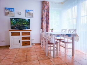 Uma área de refeições com uma mesa e cadeiras brancas, uma televisão num móvel e duas pinturas na parede.
