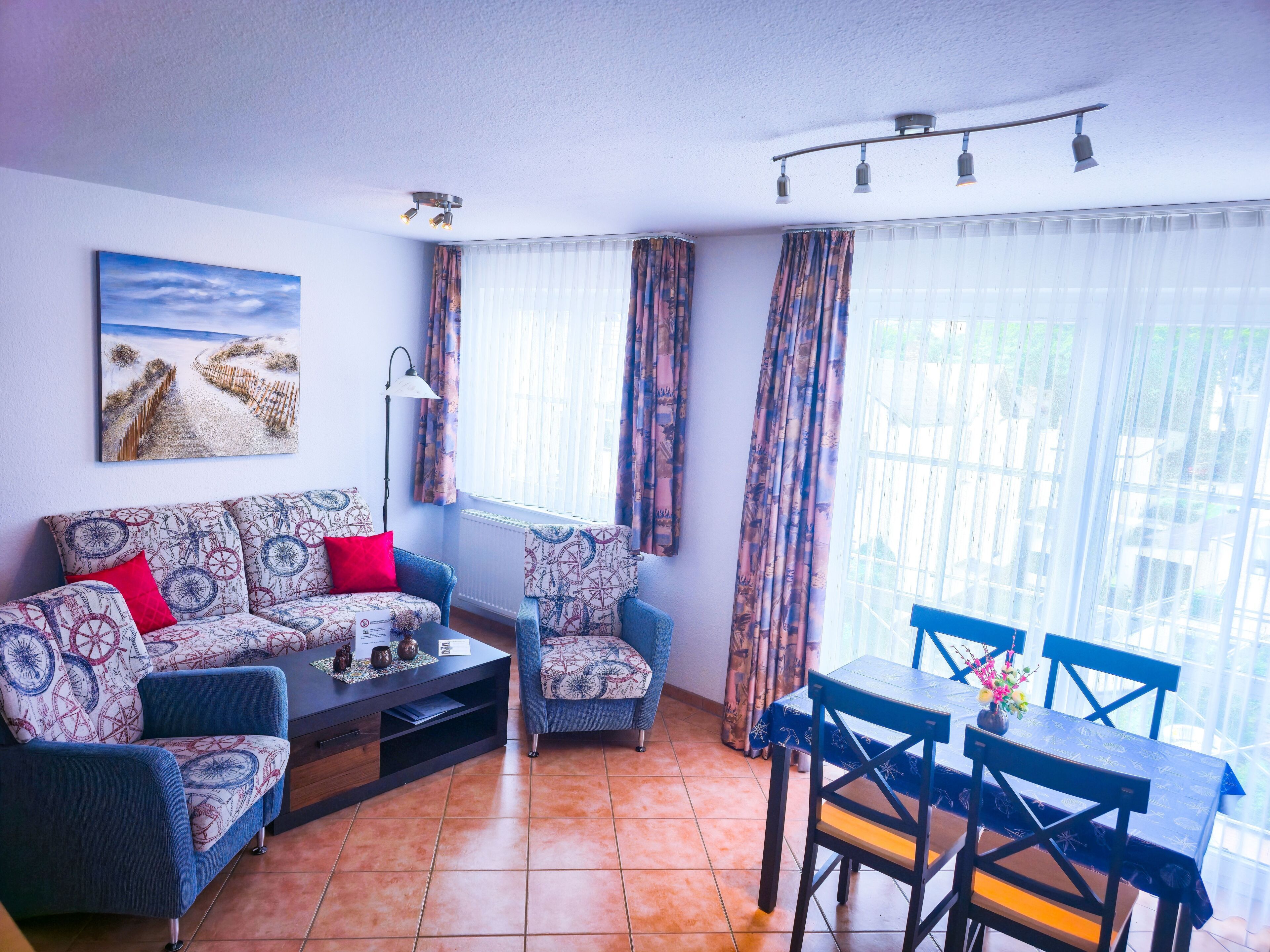 Ein Wohnzimmer mit einer Couch, einem Couchtisch und einem Esstisch mit Stühlen. An der Wand hängt ein Gemälde und es gibt ein Fenster mit Vorhängen.