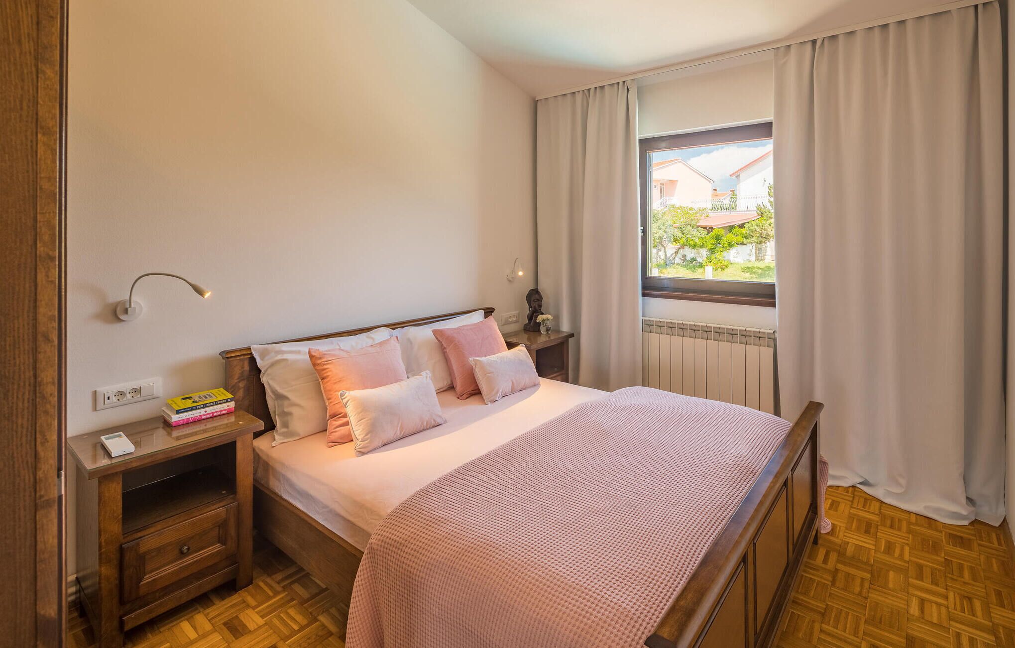 A bedroom with a wooden bed, a nightstand with a lamp, a window with curtains, and a view of greenery outside.