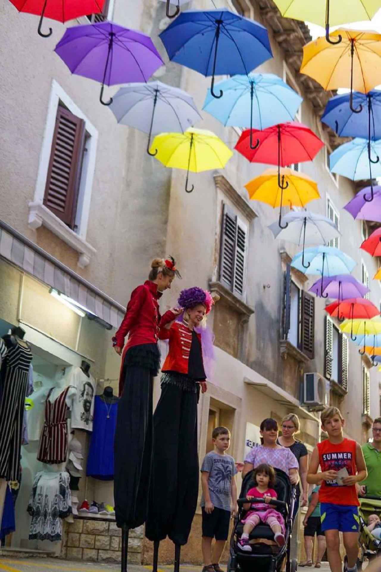 A street with colorful umbrellas hanging overhead, a group of people, and a shop with clothing items on display.