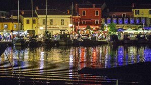 Una escena nocturna a la orilla del río con barcos amarrados, edificios con letreros iluminados y una multitud de gente.
