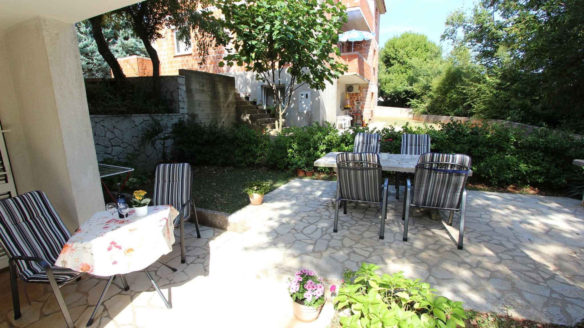 Un patio al aire libre con suelo de piedra, sillas a rayas, una mesa con mantel floral y plantas en macetas.