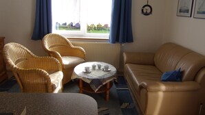 Un salon chaleureux, meublé de meubles en osier, avec un canapé en cuir et une vue sur un jardin, visible à travers une fenêtre ornée de rideaux bleus.