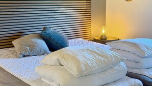 Cama bem arrumada com roupa de cama branca e várias almofadas, cabeceira de ripas de madeira, uma pequena mesa com abajur e um espelho redondo na parede.