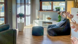 Uma sala de estar moderna com um sofá, um banquinho e uma mesa de centro. Há uma planta em vaso e uma janela com vista para o exterior.