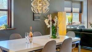 Uma sala de jantar com uma mesa branca, cadeiras cinzentas, uma garrafa de vinho e um vaso com flores.