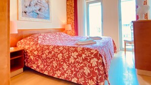 A hotel room with a bed, a nightstand, a lamp, a painting on the wall, and a view of the outside through a window. - Residence Bleichröder- Charlotte, VS Sass - Residence Bleichröder vacation apartment (Heringsdorf)