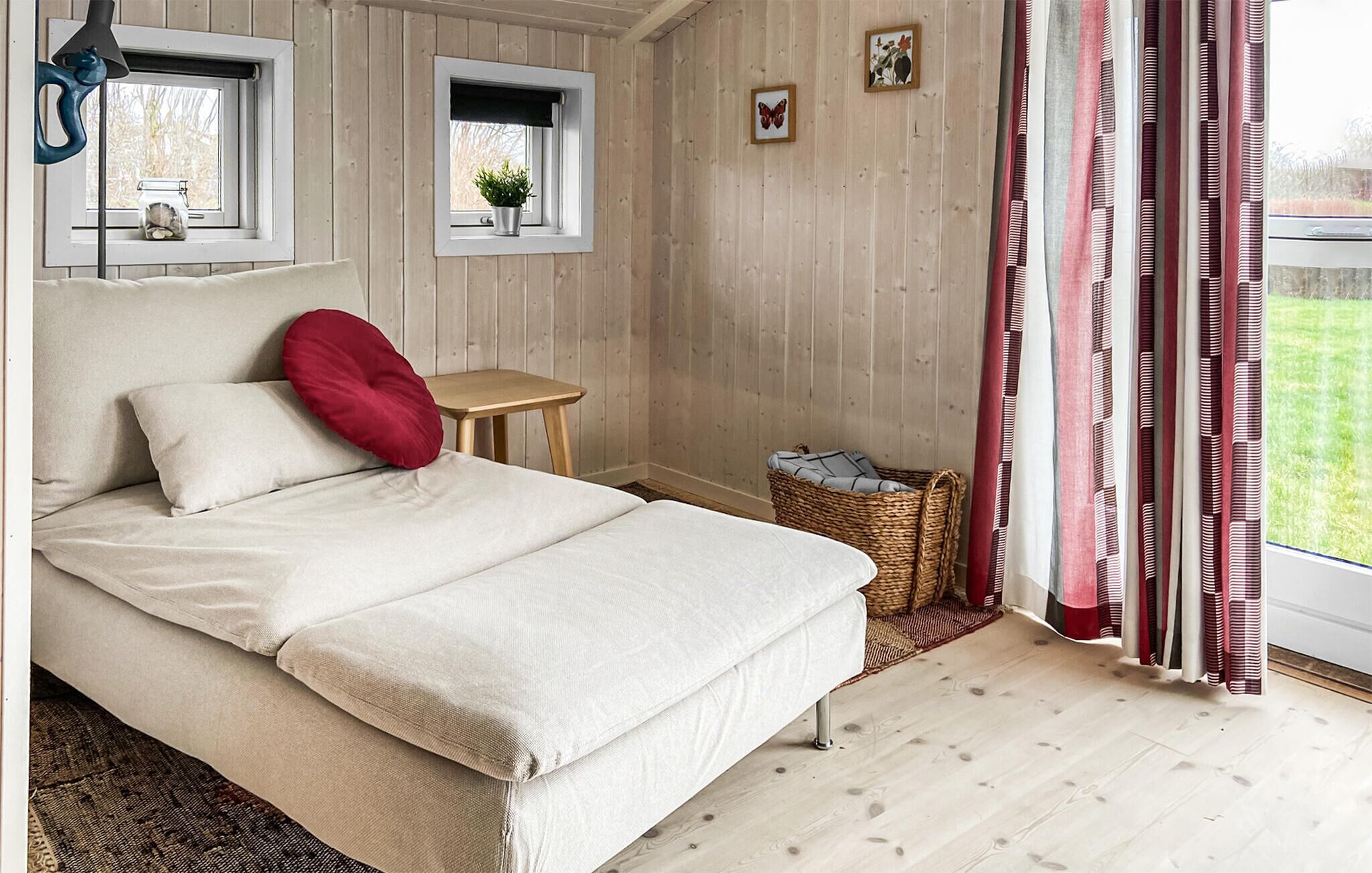 A bedroom with a bed, a small table, a window with a plant, a basket, and a curtain.